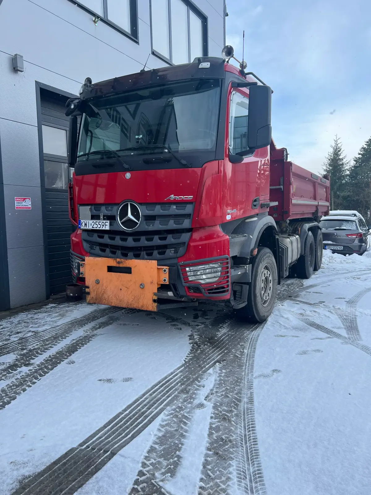 Czerwona wywrotka Mercedes-Benz Arocs z zamontowanym pługiem na zaśnieżonym placu przed halą serwisową w Tymbarku